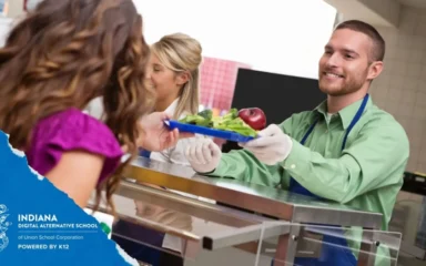 A man in a green shirt and apron smiles while handing a lunch tray with salad and an apple to a woman at a cafeteria counter. The mood is friendly and welcoming.