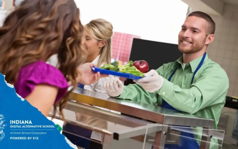 A man in a green shirt and apron smiles while handing a lunch tray with salad and an apple to a woman at a cafeteria counter. The mood is friendly and welcoming.