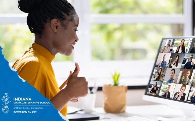A woman in a yellow shirt gives a thumbs-up to a computer screen displaying a video call with multiple participants. A logo for Indiana Digital Alternative School is visible.