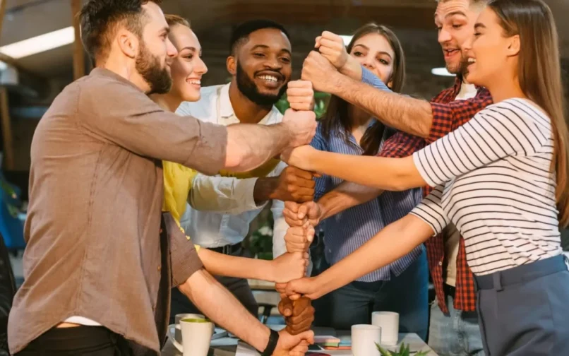 A diverse group of six people in casual attire stand in a circle, smiling and stacking their hands in unity. The scene conveys teamwork and positivity.