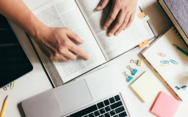 Hands point at an open book amidst a study setup with a laptop, colorful paper clips, sticky notes, and a pen, conveying focus and organization.