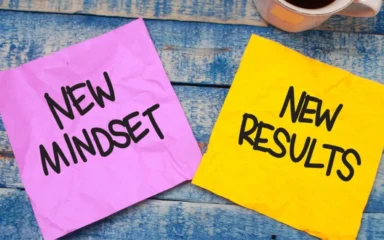 Two crumpled sticky notes on a blue wooden surface. The pink note reads "New Mindset," and the yellow reads "New Results." A cup of coffee is visible nearby.