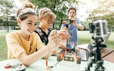 Three teens are in a park working on a robotics project. One holds wires, another films with a smartphone, and a tripod-mounted camera is nearby. They are focused and smiling.