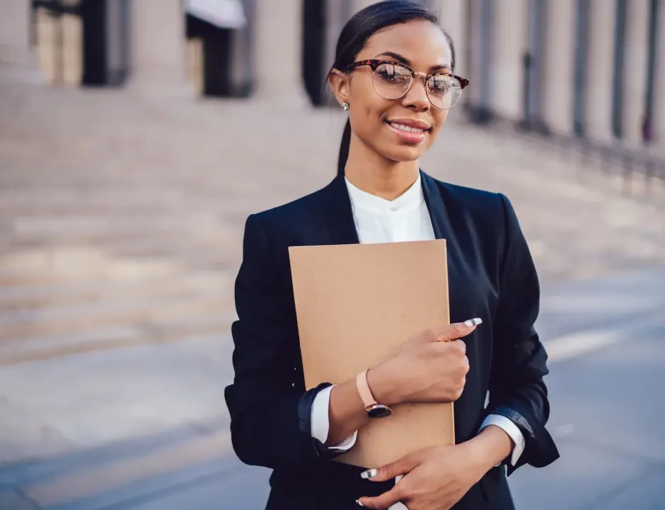 Confident woman in glasses and a black blazer holds a file folder, standing on steps outdoors. The setting feels professional and empowering.