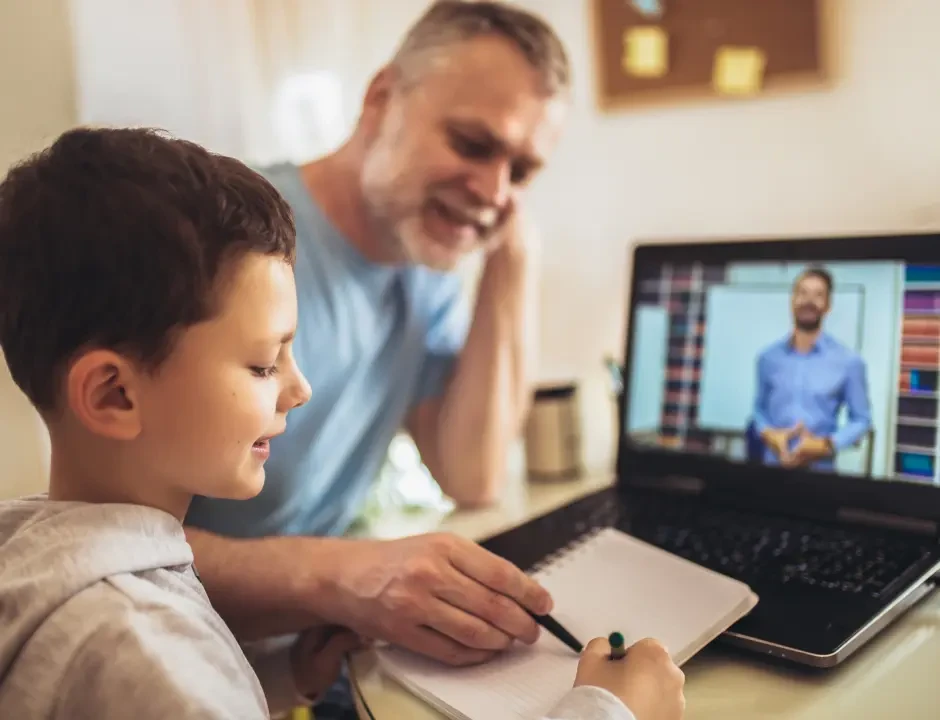 A young boy writes in a notebook while a man, possibly his father, attentively watches. A laptop displays an online teacher. The mood is focused and supportive.