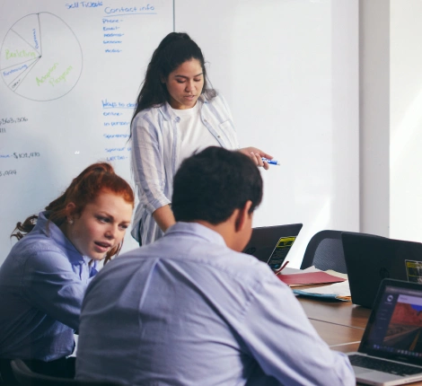 A group of three people collaborate in an office. Two are seated with laptops, discussing, while one stands near a whiteboard, holding a marker. The atmosphere is focused.