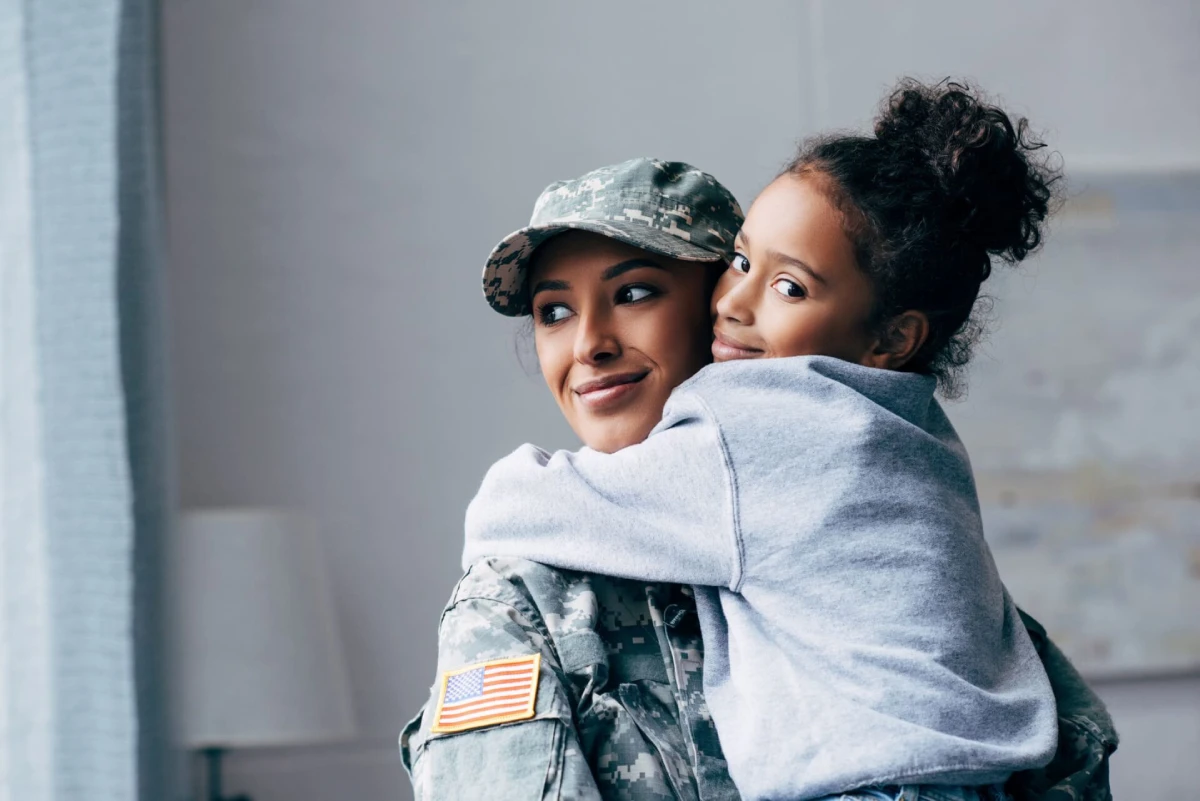 A smiling woman in military uniform holds a young girl who is hugging her. The girl wears a gray sweatshirt, conveying warmth and affection.